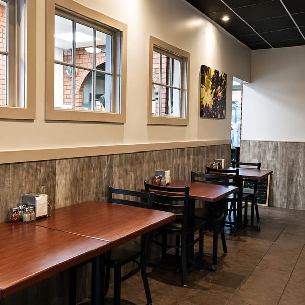 Empty restaurant dining room with dark wood tables, black chairs, and windows looking out to brick.